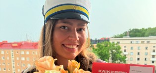 Ylioppilaslakkiin pukeutunut Matilda Gustafsson hymyilee kameralle. Hänellä on sylissään keltaisia ruusuja sekä TAIn punainen kansio. Taustalla näkyy kerrostaloja.