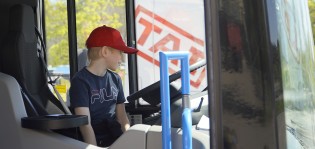 Punaiseen lippikseen pukeutunut alakoululainen istuu bussinkuljettajan paikalla ja ihmettelee ohjaamoa. Taustalla näkyy rekan ohjaamo, jossa on valkoisella pohjalla punainen TAI-logo.
